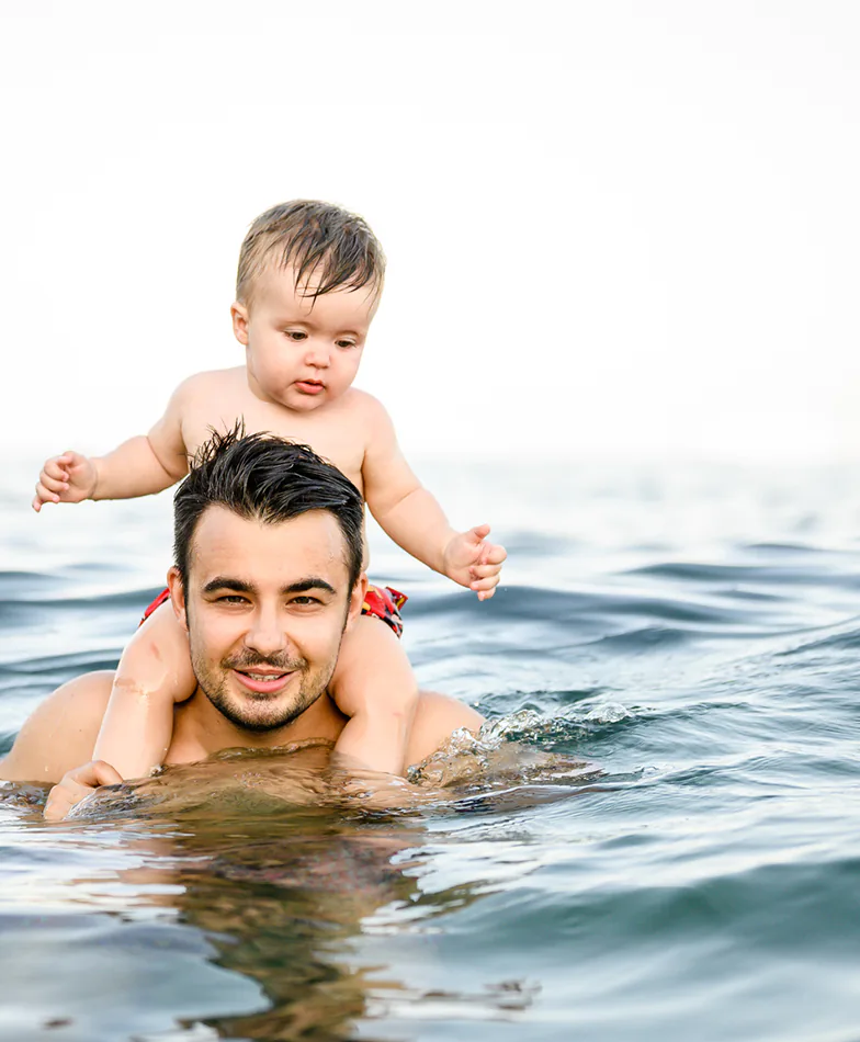 Father neck deep in water with his baby son sitting on his shoulders.